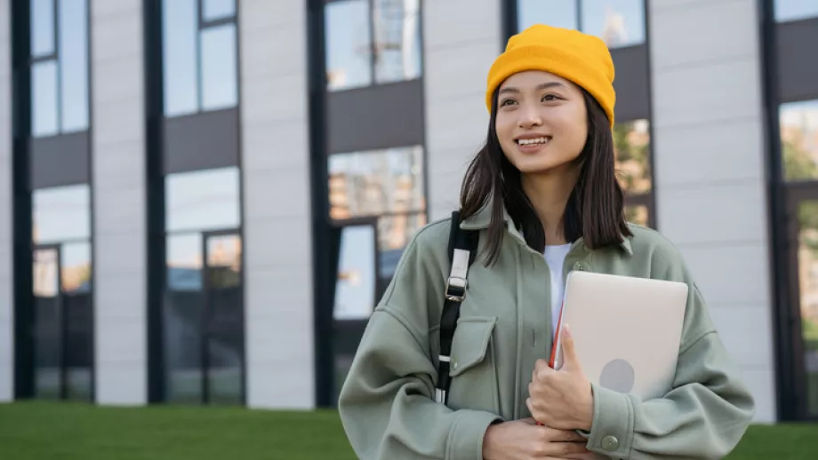 Student in yellow hat carries laptop in front of university